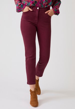 Persona con pantaloni aderenti bordeaux a vita alta, camicia a maniche lunghe con fantasia floreale e stivaletti in camoscio marrone, in piedi con le mani in tasca.