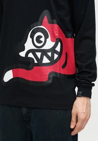 Μαύρο μακρυμάνικο πουκάμισο με τολμηρό κόκκινο, λευκό και μαύρο γραφικό μιας στυλιζαρισμένης καρτούν κεφαλής σκύλου με αιχμηρά δόντια και μεγάλο στρογγυλό μάτι.