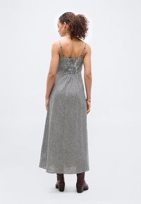 Γυναίκα με σγουρά μαλλιά, φορώντας ασπρόμαυρο φόρεμα με καρό χωρίς μανίκια και καφέ μποτάκια μέχρι τον αστράγαλο, στέκεται με την πλάτη προς τα εμπρός σε απλό φόντο.