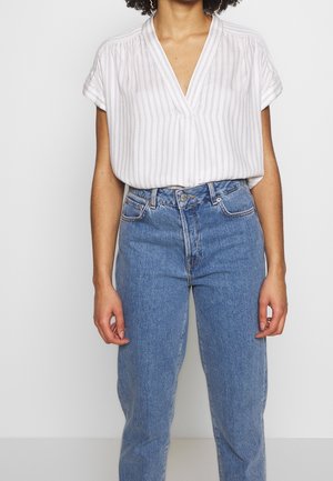 Kvinde iført hvid kortærmet V-halsstribet bluse, stoppet ned i højtaljede blå jeans, stående mod en ensfarvet baggrund.