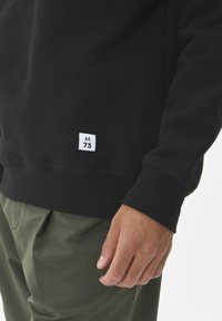 Zwarte sweatshirt met een geribbelde kraag en mouwen, voorzien van een wit label met "M 73" onderaan, gecombineerd met olijfgroene broek.