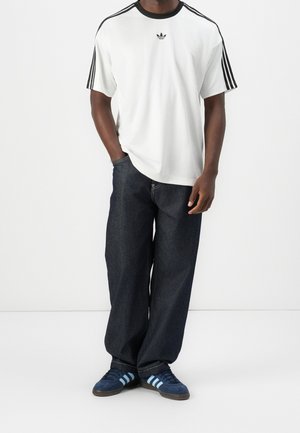 Mann trägt ein weißes Adidas Kurzarmshirt mit schwarzen Streifen, dunkle lockere Jeans und blaue Adidas Sneakers mit hellblauen Streifen.