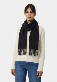 Bufanda negra hecha de material suave, con extremos desflecados. Se lleva colocada alrededor del cuello sobre un suéter ligero y jeans.