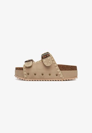 Beige slide sandals with two adjustable straps, decorative stitching, and gold-tone hardware. Cork footbed and textured sole with studs.