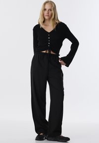 Zwarte cropped sweater met een V-hals en vetersluiting met de tekst "TWIST." Gecombineerd met losse zwarte broeken en zwarte schoenen.