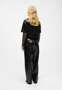 Personne blonde portant un haut noir brillant avec un motif en serpent à manches courtes et un pantalon large assorti, se tenant devant un fond uni.