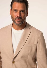 Blazer beige avec un tissu texturé, fermeture à un bouton, revers crantés et poche poitrine, porté sur un T-shirt blanc.