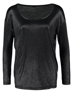Kvinde iført en skinnende sort langærmet top, mørke slanke jeans, sorte ankelstøvler, bærer en stor sort håndtaske med nitter.