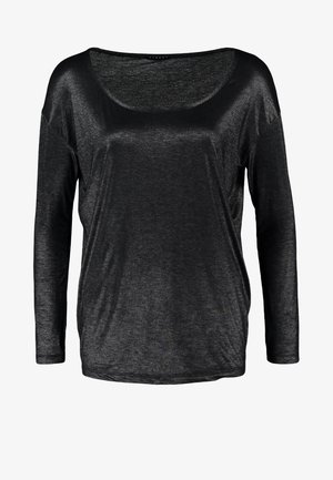 Kvinde iført en skinnende sort langærmet top, mørke slanke jeans, sorte ankelstøvler, bærer en stor sort håndtaske med nitter.