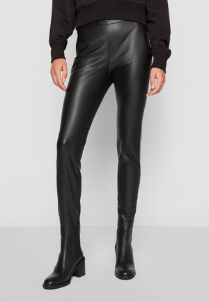 Femme portant des leggings noirs en simili cuir associés à des bottines à talons noires et un pull noir, debout devant un fond uni.