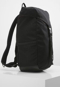 Mochila negra con una tela texturizada, correas acolchadas y un cierre de gancho y bucle. Cuenta con un bolsillo lateral y herrajes de plástico negro.