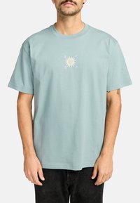 Hellblaues Baumwoll-T-Shirt, kurze Ärmel, Rundhalsausschnitt, mit einem zentralen mehrfarbigen Sonnenmotiv und dem Text "RVCA" darum herum.