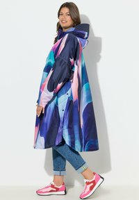 Langärmeliger Regenmantel in Marineblau mit buntem abstraktem Muster, mit Kapuze und Druckknöpfen. Kombiniert mit umgeschlagenen Jeans und pinken Sportschuhen.