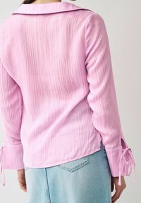 Blouse rose clair à rayures avec un col et des accents en nœud sur les manches. Tissu doux avec une texture fluide, superposé à un bas en denim.