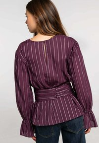 Blusa morada a rayas con mangas largas, cintura peplum y detalle de botón en la espalda. La tela tiene una textura suave y una cintura ajustada.
