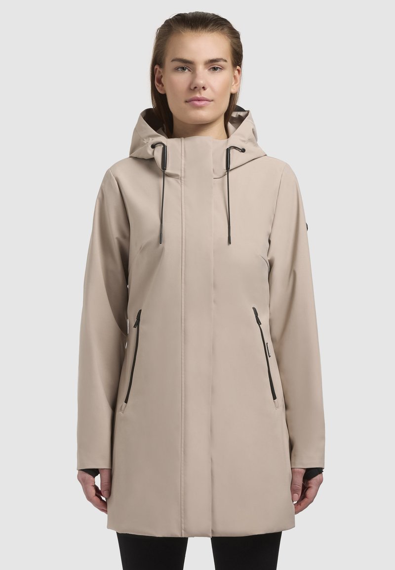 Beige vandtæt jakke med hætte, frontlynlås og sorte lynlåsdetaljer. Har lange ærmer og sidelommer. Glat tekstur.