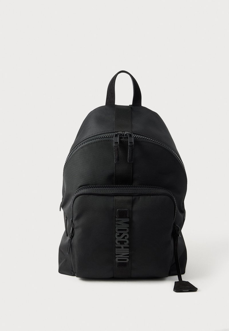 Μαύρο σακίδιο Moschino με πάνω λαβή, μπροστινή τσέπη με φερμουάρ και ορατό λογότυπο στο λουράκι της μπροστινής τσέπης σε απλό φωτεινό φόντο.