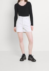 Musta pitkähihaisen paita, jossa pitsikaulus, yhdistettynä valkoisiin korkeavyötäröisiin shortseihin. Malli käyttää mustia urheilukenkiä, joissa on violetteja yksityiskohtia.