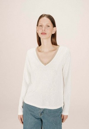 Haut à manches longues blanc avec un décolleté en V, orné d'une bordure beige décorative. Fabriqué en tissu doux et texturé, associé à un jean bleu clair.