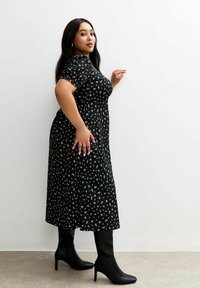 Vestido midi negro con mangas cortas, que presenta un estampado floral. Combinado con botas negras ajustadas hasta el tobillo, de pie contra un fondo neutro.