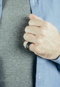 Fekete, textúrált gyűrű egy kézen, szürke póló és világoskék gombos ing háttérrel. Figyelj a gyűrű spirális mintázatára.