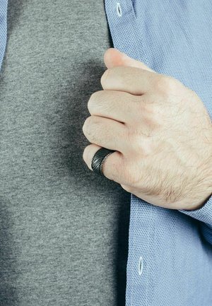 Fekete, textúrált gyűrű egy kézen, szürke póló és világoskék gombos ing háttérrel. Figyelj a gyűrű spirális mintázatára.