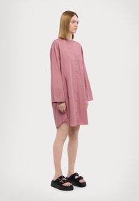 Abito camicia oversize rosa con maniche lunghe, una tasca frontale e vestibilità rilassata. Abbinato a sandali con plateau neri.