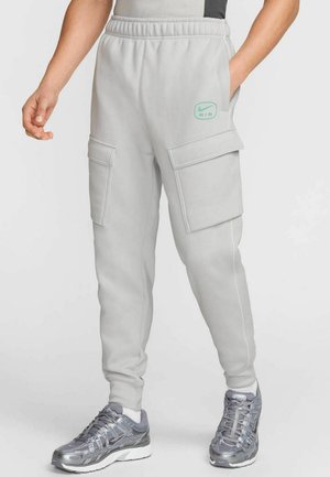Pantalones cargo de color gris claro con cintura elástica, dos bolsillos laterales y costuras blancas en contraste. Presenta un pequeño logo de Nike en color teal en el muslo.