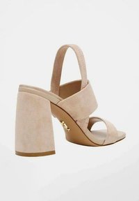 Sandal con tacco alto in suede beige, con tacco a blocco largo, cinturino alla caviglia regolabile e design incrociato sulla parte anteriore del piede. Accenti in tono oro visibili.