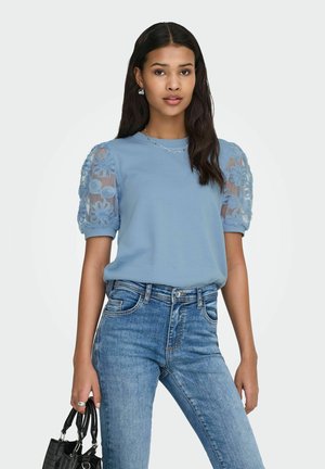 Vrouw draagt lichtblauwe blouse met bloemenkant-pofmouwen en blauwe jeans, houdt een zwarte handtas langs haar zij tegen een witte achtergrond.