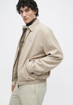 Beige bomberjack met een rits aan de voorkant, elastische manchetten en tailleband. Kenmerkt geborduurd logo op de borst. Gemaakt over een bruine coltrui.