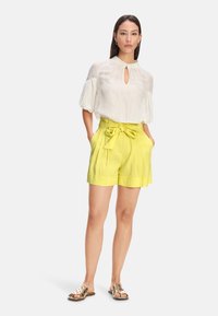 Weiße Bluse mit einem Schlüssellochdetail und puffartigen Ärmeln, kombiniert mit leuchtend gelben hochgeschnittenen Shorts mit einem Bindegürtel. Sandalen mit goldenen Akzenten.