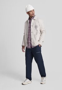 Giacca beige con logo, camicia a quadretti, pantaloni blu navy e sneakers bianche. Un berretto di lana completa l'outfit. Look casual a strati.