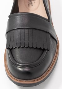 Mocassin en cuir noir avec détail à franges sur la tige, bout arrondi et semelle en caoutchouc beige avec surpiqûres décoratives.