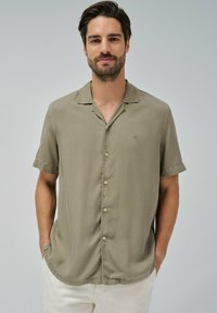 Camisa de botones de manga corta en tejido ligero color caqui. Cuenta con cuello, botones frontales y un logotipo sutil en el pecho. Textura suave.