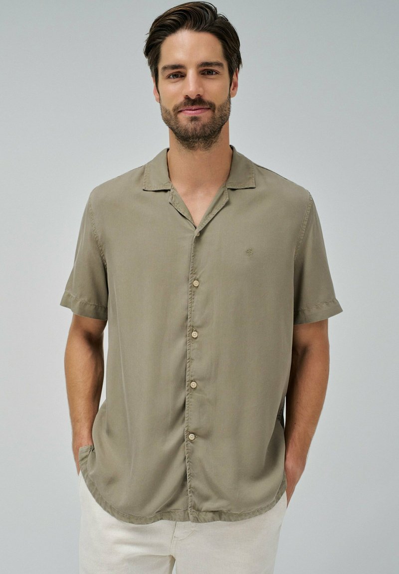 Camisa de botones de manga corta en tejido ligero color caqui. Cuenta con cuello, botones frontales y un logotipo sutil en el pecho. Textura suave.
