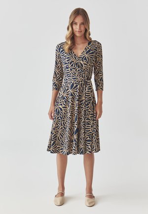Marine jurk met beige bladerenpatroon, V-hals, driekwartmouwen, taille die is aangetrokken, en een uitlopende rok, gedragen met beige balletschoenen.