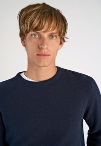 Marineblauer, strukturierter Pullover mit rundem Halsausschnitt, getragen über einem weißen Hemd. Der Stoff weist ein dezentes Muster und gerippte Bündchen auf.