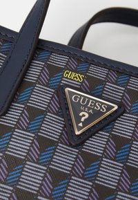 Primo piano di una borsa con motivo blu e viola, con manici blu navy e una targhetta triangolare con il logo Guess USA.
