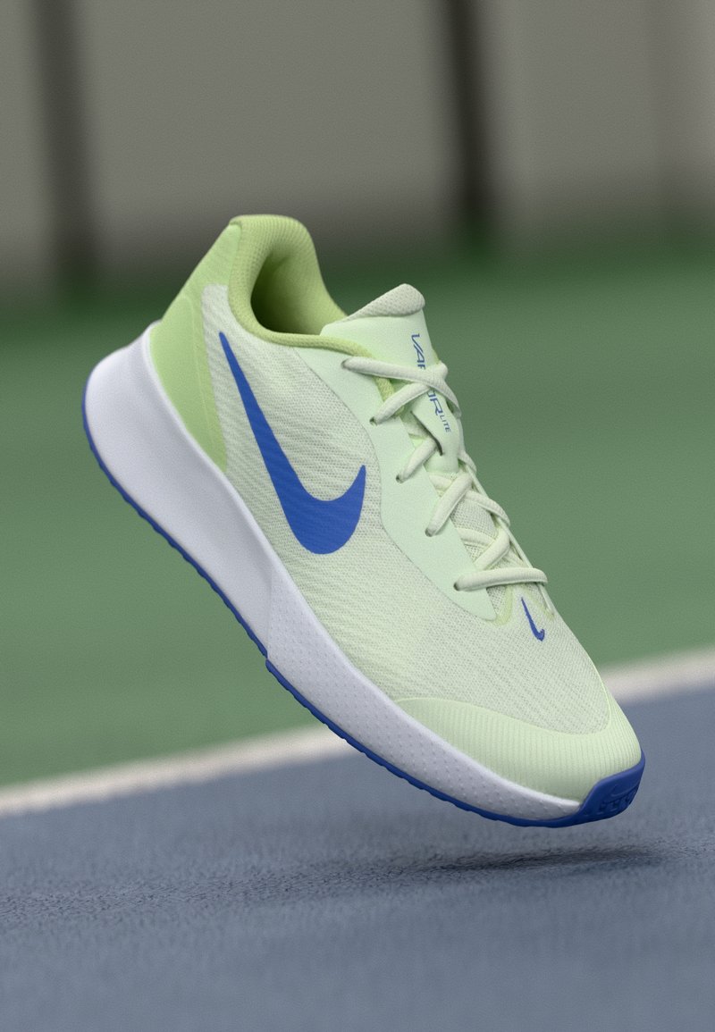 Hellgrüner und weißer Sportschuh mit blauem Nike-Swoosh, weißen Schnürsenkeln und blauer Sohle auf einer grünen und blauen Sportoberfläche.