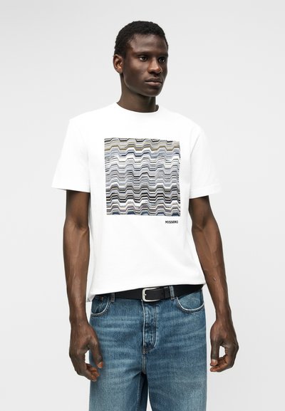 Άνδρας που φοράει λευκό T-shirt με πολύχρωμο ζιγκ-ζαγκ σχέδιο τετραγώνου και το κείμενο "MISSONI", σε συνδυασμό με μπλε τζιν και μαύρη ζώνη.