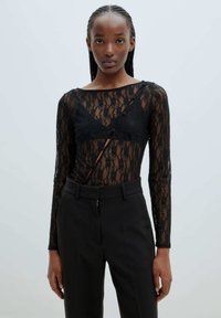 Schwarzes Spitzenoberteil mit langen Ärmeln, das ein transparentes Design und Knopfakzente aufweist. Kombiniert mit maßgeschneiderten schwarzen Hosen für einen eleganten Look.