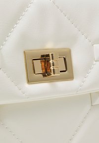 Vit quiltad läderhandväska i närbild med en glänsande guldlåsning och vit sömnad, som framhäver en slät textur och geometriskt mönster.