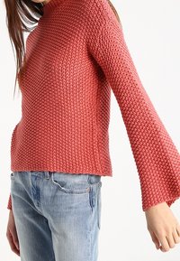 Eine Frau trägt einen strukturierten, langärmeligen, gestrickten rosa Pullover mit ausgestellten Ärmeln und hellblauen Jeans vor einem weißen Hintergrund.