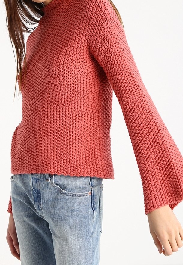 Eine Frau trägt einen strukturierten, langärmeligen, gestrickten rosa Pullover mit ausgestellten Ärmeln und hellblauen Jeans vor einem weißen Hintergrund.