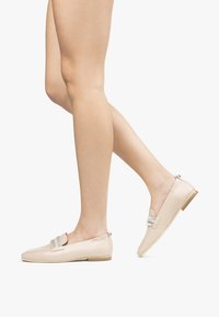 Beige læder loafers med en glat tekstur, udstyret med et dekorativt sølvfarvet accentbånd og en lav træhæl.