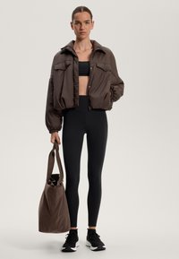 Ruskea puhvijakku, jossa on suuret etutaskut, yhdistetty mustaan crop-toppiin ja korkeavyötäröisiin legginsseihin, pitää mukanaan yhteensopivaa olkalaukkua.