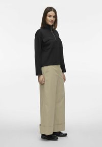 Sort zip-up trøje med to brystlommer, parret med beigefarvede brede bukser. Begge beklædningsgenstande har en glat tekstur og en afslappet pasform.
