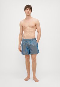 Blå svømmeshorts laget av et lett, glinsende materiale, med elastisk midjebånd og sidelommer. Modell står barbeint.