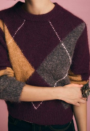 Personne portant un pull en maille duveteuse à blocs de couleurs violet, moutarde et gris, manches retroussées, debout devant un fond rose.